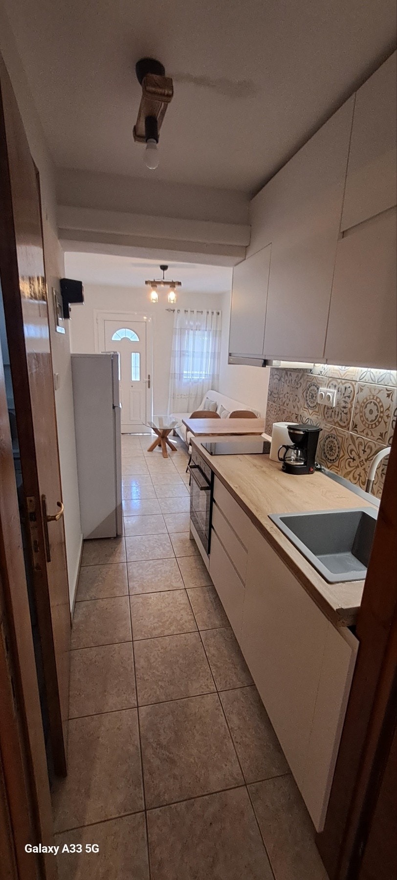 A compact kitchen area is visible, featuring modern cabinetry and tiled walls. A refrigerator stands at one end, while a sink is integrated into the countertop. Natural light is welcomed through the doorway at the far end, leading to a cozy living space.