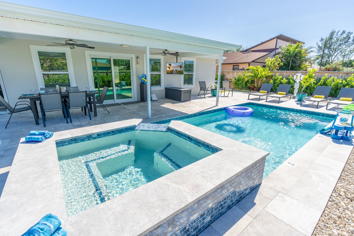 A private backyard features a saltwater pool alongside a spa, both surrounded by a tiled patio. Multiple lounge chairs are positioned by the pool, while a dining table is set for outdoor meals. Lush greenery provides a sense of privacy.