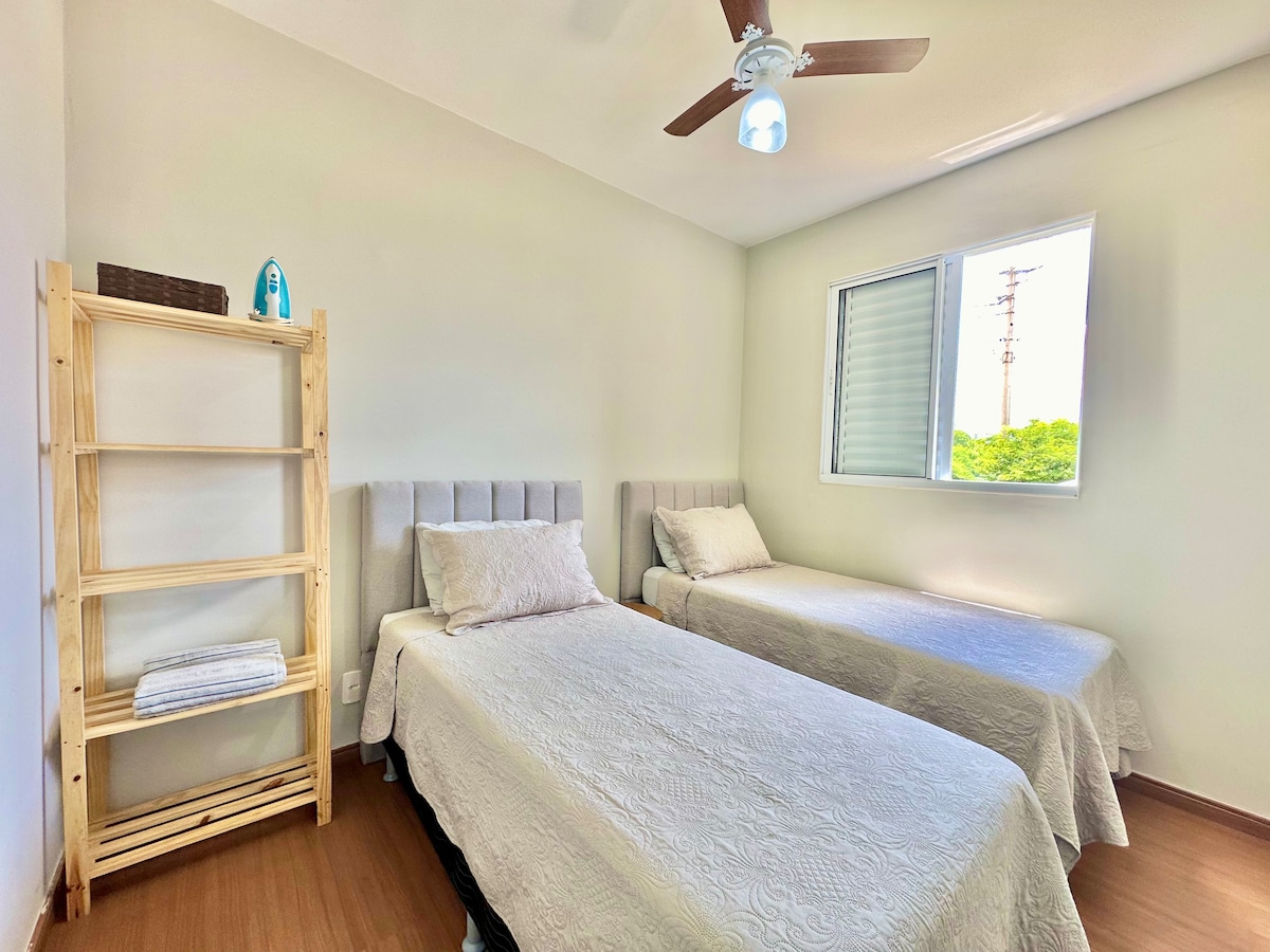A bedroom featuring two single beds with light-colored linens and upholstered headboards. A wooden shelf stands beside the beds, displaying a neatly rolled towel. Natural light enters through a window, creating a bright and airy ambiance.