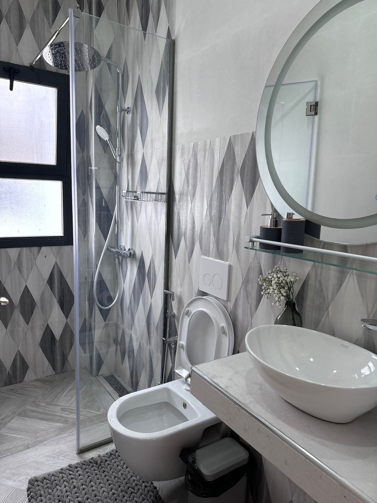 The modern bathroom features a walk-in shower with glass panels, accompanied by a circular mirror above a stylish basin. A white toilet is positioned next to a small wastebasket, while a decorative plant adds a touch of elegance to the countertop. Natural light filters through a window.