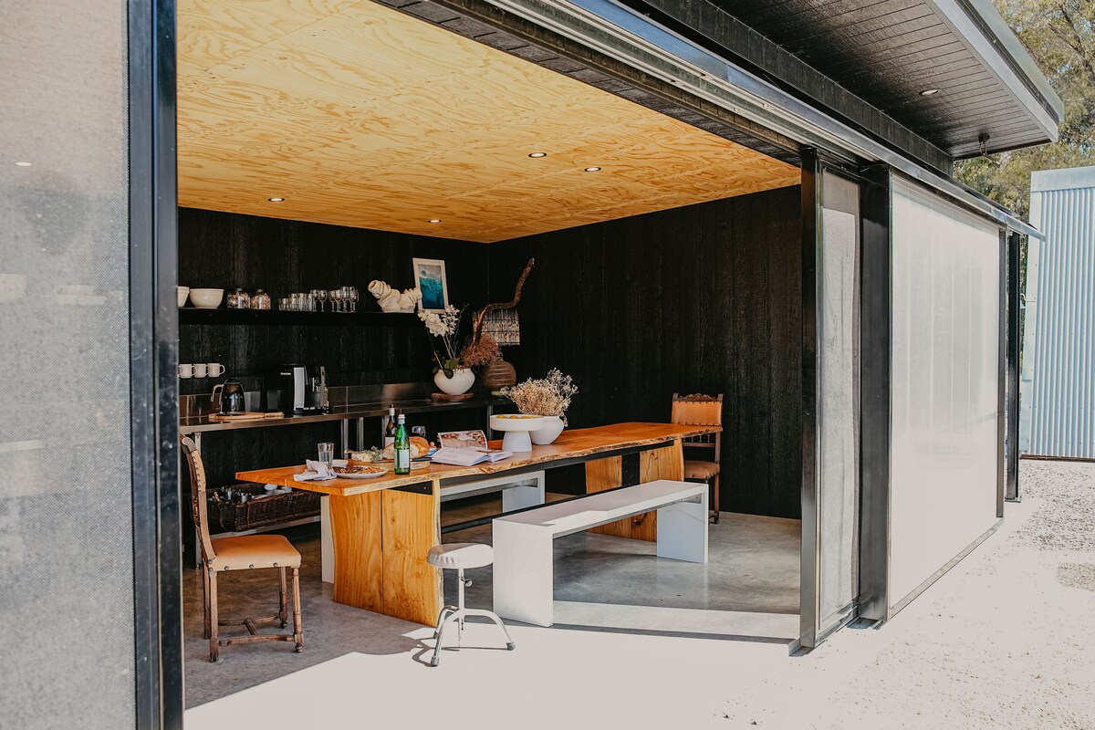 An outdoor dining area is showcased under a wooden ceiling, featuring a long wooden table surrounded by a mix of chairs. A well-equipped kitchen space is visible in the background, complete with shelves displaying glassware and decorative items.