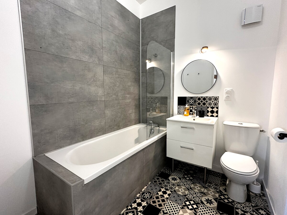 A modern bathroom features a bathtub set against textured gray tiles. A sleek white vanity with an oval mirror is positioned nearby, providing storage space. Decorative black and white patterned tiles enhance the floor, while a window ensures natural light fills the room.