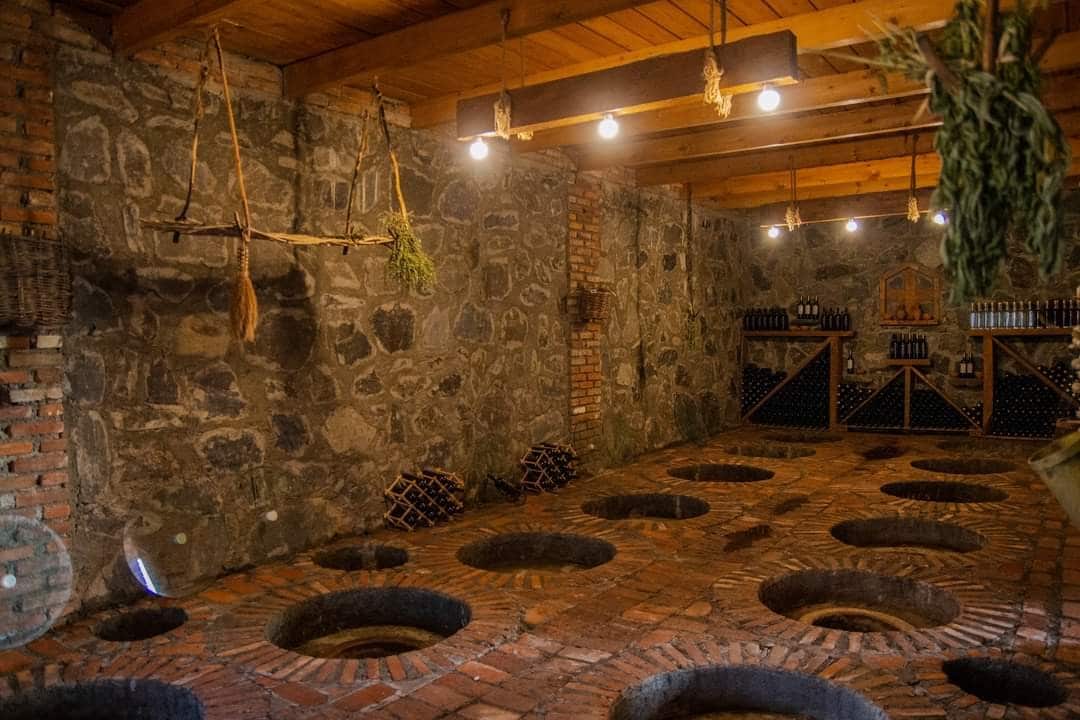 A rustic cellar features stone walls and a wooden ceiling supported by exposed beams. Circular openings in the brick floor are arranged neatly, and shelves lined with bottles are visible against one wall. Soft lighting highlights the natural textures of the space.