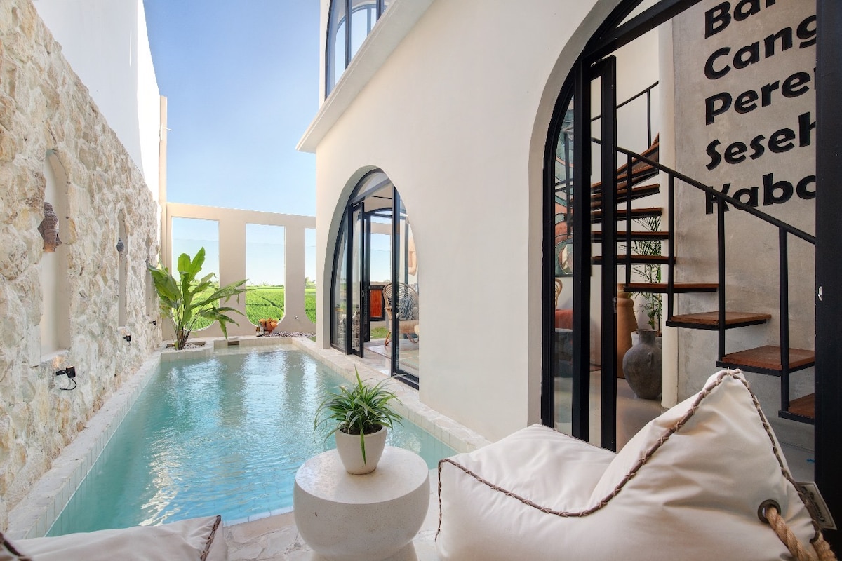 An outdoor area features a private pool surrounded by natural stone walls and large arched windows. A small white table stands nearby, complemented by plush seating adorned with light-colored cushions. Lush greenery is visible, enhancing the tranquil ambiance of the space.