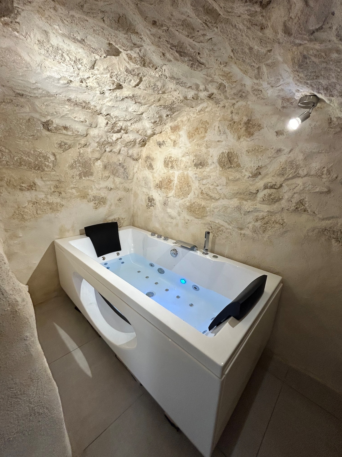 A modern bathtub with built-in jets is situated in a stone-walled alcove, illuminated by a soft overhead light. The soothing blue water is accented by small lights, creating a serene setting for relaxation.