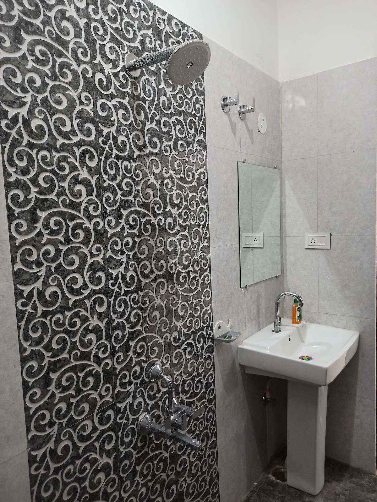 A modern bathroom features a patterned gray wall with a rainfall showerhead and an additional hand-held shower option. A compact white sink is mounted on the wall, accompanied by a mirror, while toiletries are neatly placed nearby, offering functional amenities.