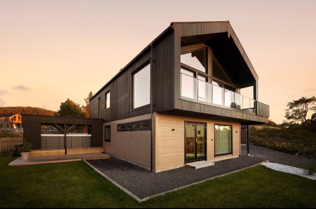A contemporary house features a striking architectural design with a high-pitched roof and large glass windows. A spacious deck extends from the second floor, offering panoramic views. The exterior showcases a mix of dark and light materials, surrounded by a well-maintained grassy area.
