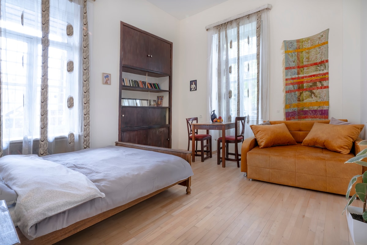 The spacious room includes a double bed with a light grey cover on a wooden frame, complemented by a comfortable sofa. Natural light filters through large windows, illuminating a small dining table accompanied by two chairs. Decorative wall art adds a touch of character.
