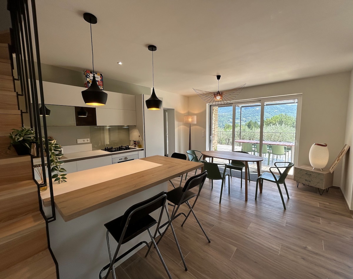 An open kitchen and living area features sleek cabinetry and a spacious wooden dining table surrounded by chairs. Large glass doors provide a view of the outside, while light fixtures illuminate the room. A staircase leads to the upper level, enhancing the inviting design.