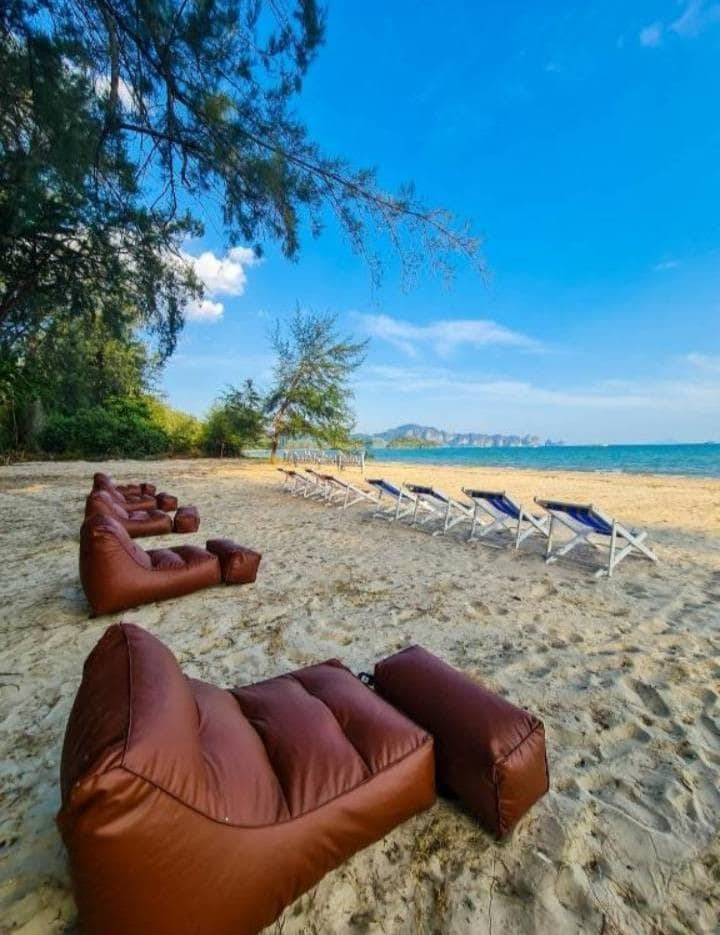 A sandy beach area is depicted with several brown lounge chairs arranged in a relaxed setting. Behind the lounge chairs, white beach loungers are lined up, while trees provide a touch of greenery in the background. The calm sea is visible, reflecting a clear blue sky.