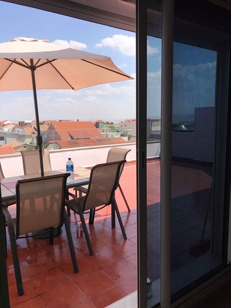 A sunlit terrace is visible through a glass door, showcasing a dining table with several chairs arranged beneath a large umbrella. The red-tiled floor reflects the sunny weather, while rooftops of nearby buildings can be seen in the background.