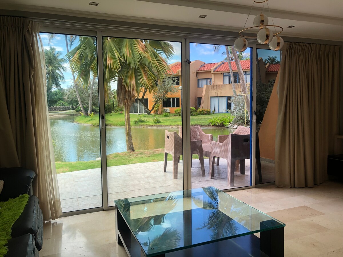 A spacious living area features large glass doors opening to a serene outdoor view. A small terrace with chairs overlooks a tranquil body of water surrounded by lush greenery and palm trees, inviting a sense of relaxation.