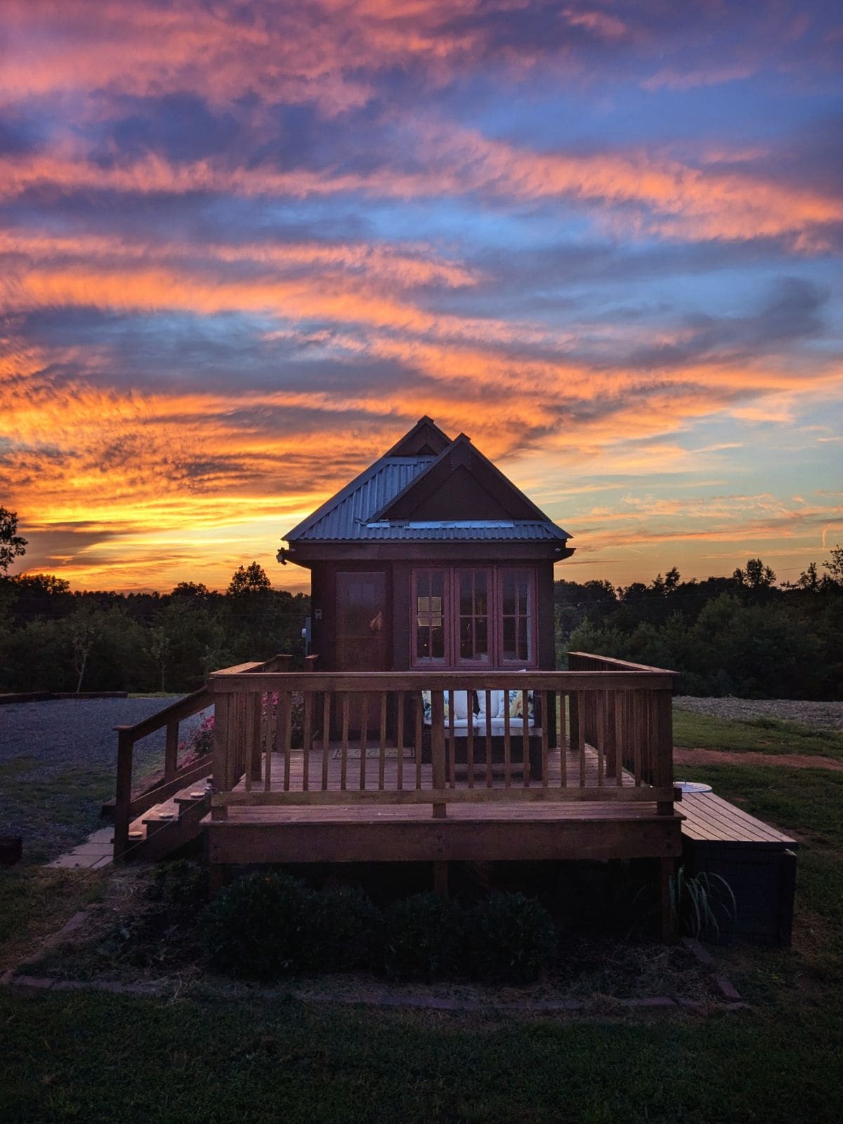 A tiny house is set against a vibrant sunset, displaying a stunning array of colors in the sky. The front porch features wooden railings and a view of the surrounding landscape, creating an inviting space for relaxation.