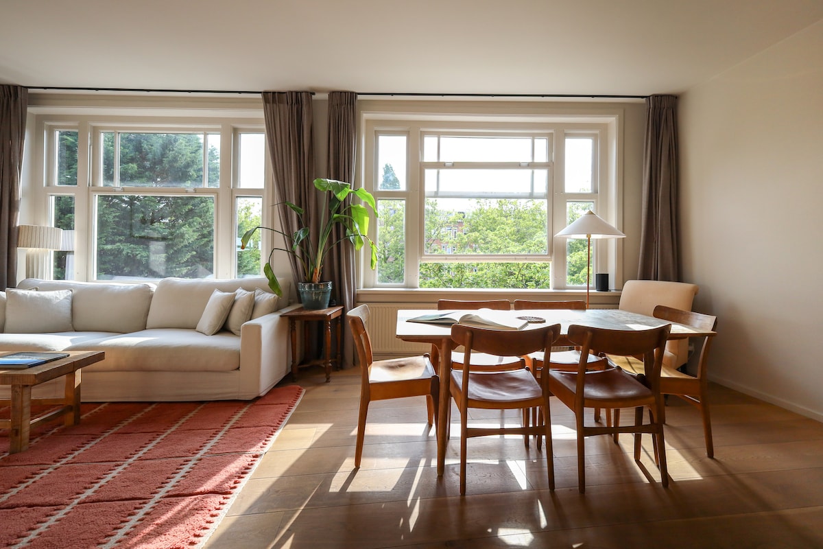 The living room features large windows that allow natural light to fill the space. A comfortable sofa rests against the wall, accompanied by a dining table and four wooden chairs. A floor lamp and a potted plant add a touch of greenery to the room.