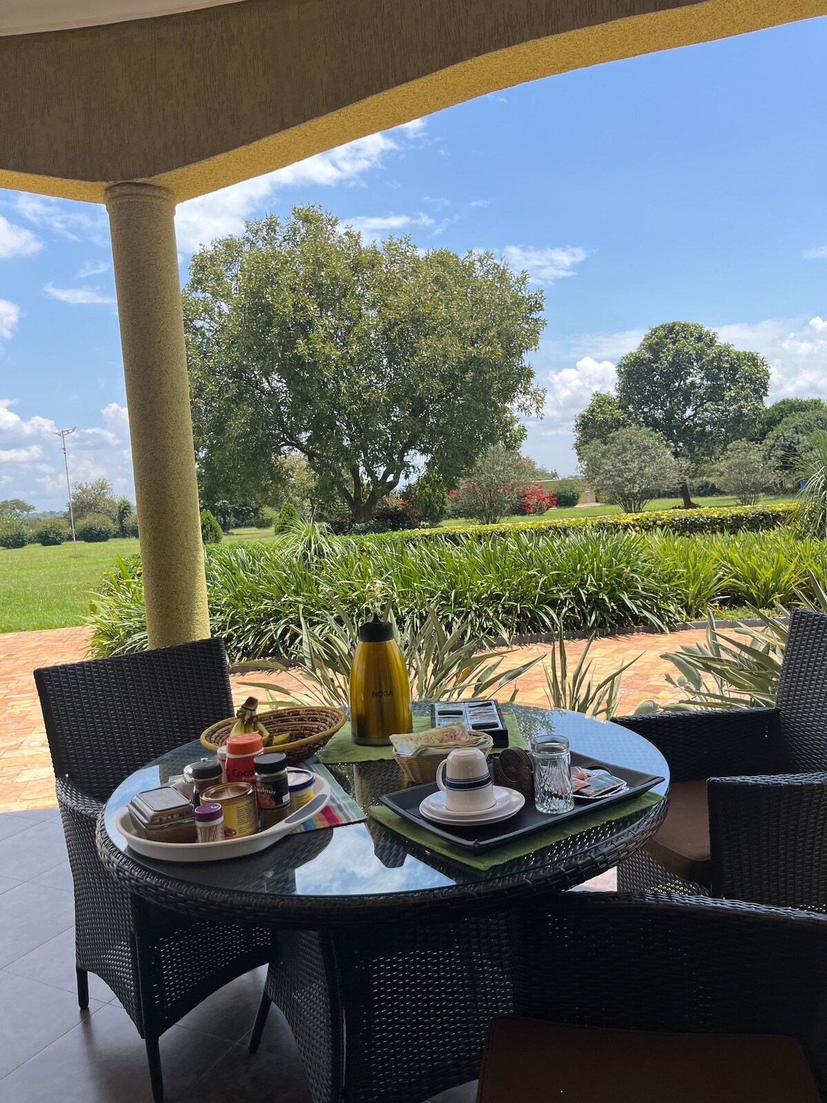 A round table is set outdoors with breakfast items, including pastries and drinks, against a backdrop of green lawns and trees. The scene is bright and airy, with blue skies and scattered clouds, offering a sense of tranquility.