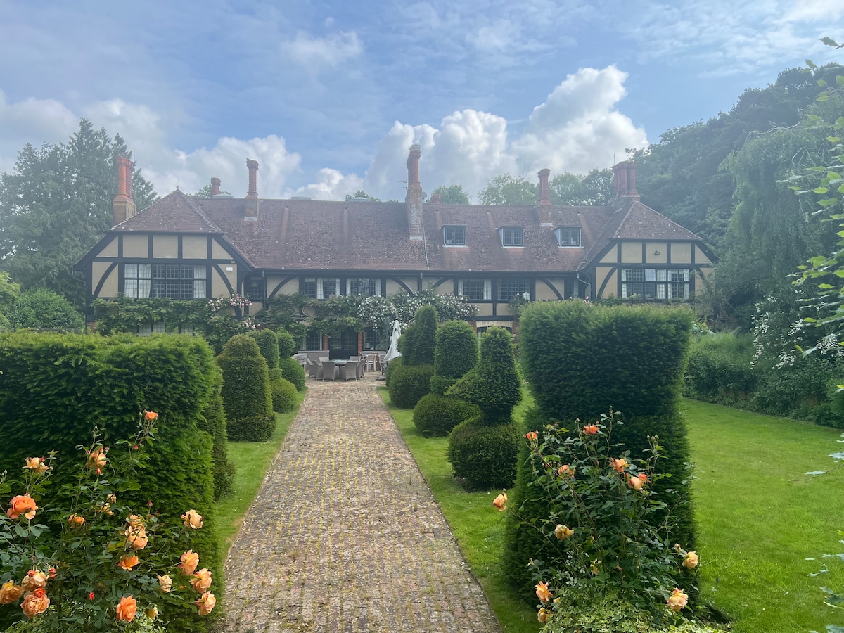 The historic house is framed by manicured hedges and vibrant rose bushes. A stone pathway leads towards the entrance, with a patio area visible for outdoor seating. The architecture reflects traditional craftsmanship, showcasing distinctive features like gabled roofs and decorative timber framing.