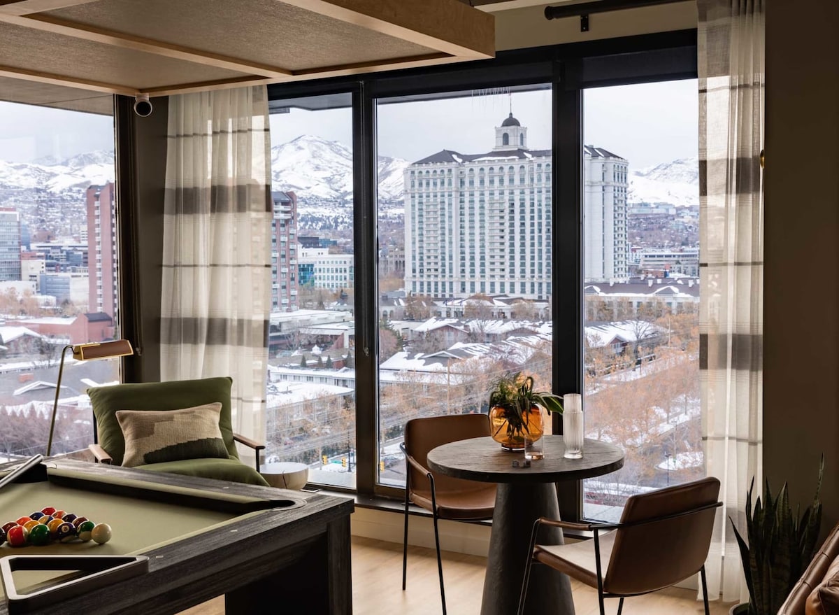 A spacious lounge area features a modern pool table beside a round dining table adorned with a decorative centerpiece. Large windows offer panoramic views of the snow-covered mountains and cityscape, allowing natural light to fill the space.