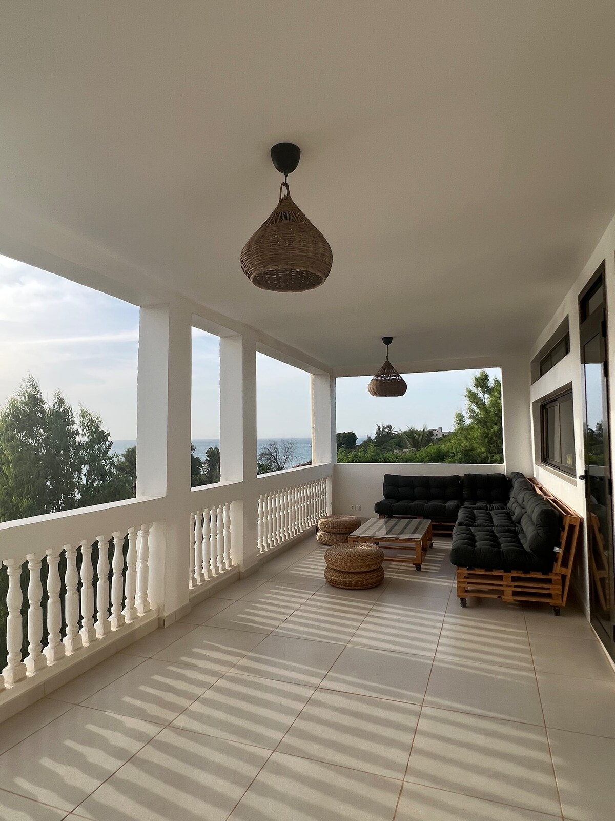 A spacious balcony offers a comfortable seating area with dark cushioned sofas and natural wooden elements. Light fixtures hang from the ceiling, and the view includes lush greenery and the sea beyond. Soft shadows are cast across the tiled floor, enhancing the serene atmosphere.