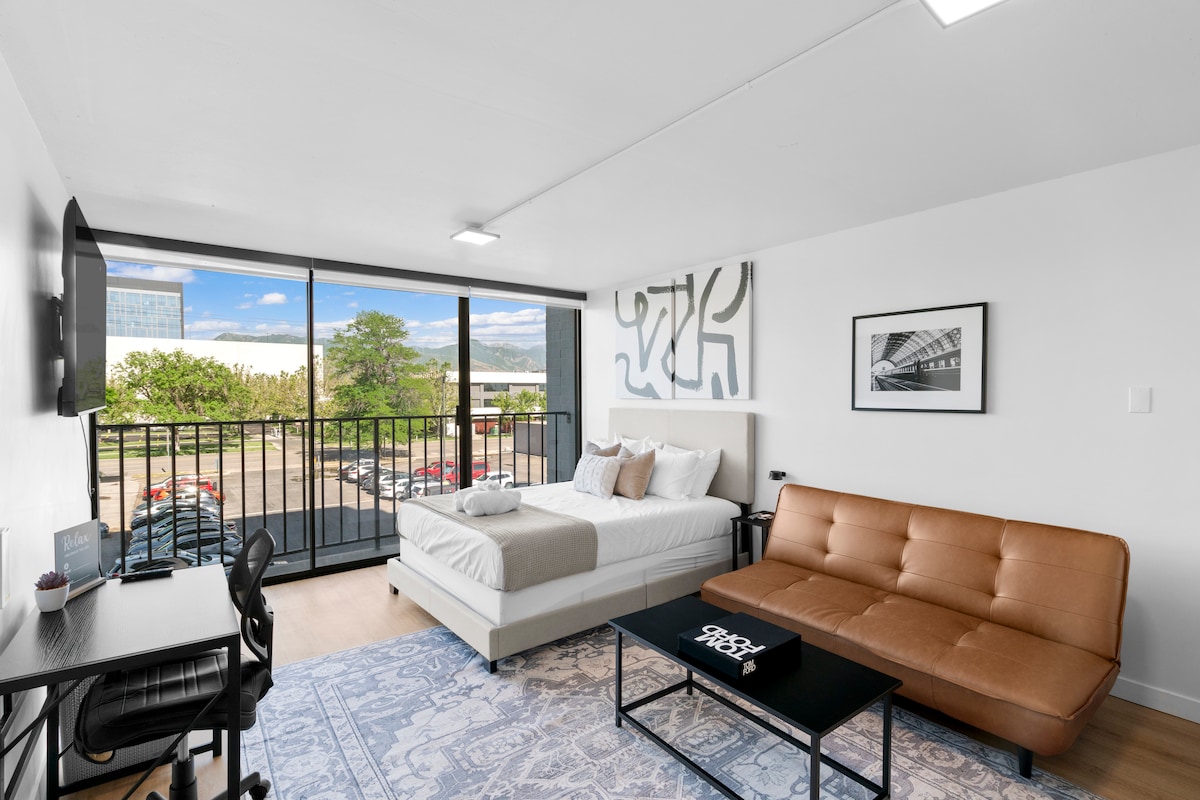 A well-appointed studio space is captured, featuring a spacious queen-sized bed and a full-sized futon. A modern desk is positioned opposite a large window, providing natural light and outdoor views. Decorative artwork enhances the room's appeal, complemented by a stylish area rug.