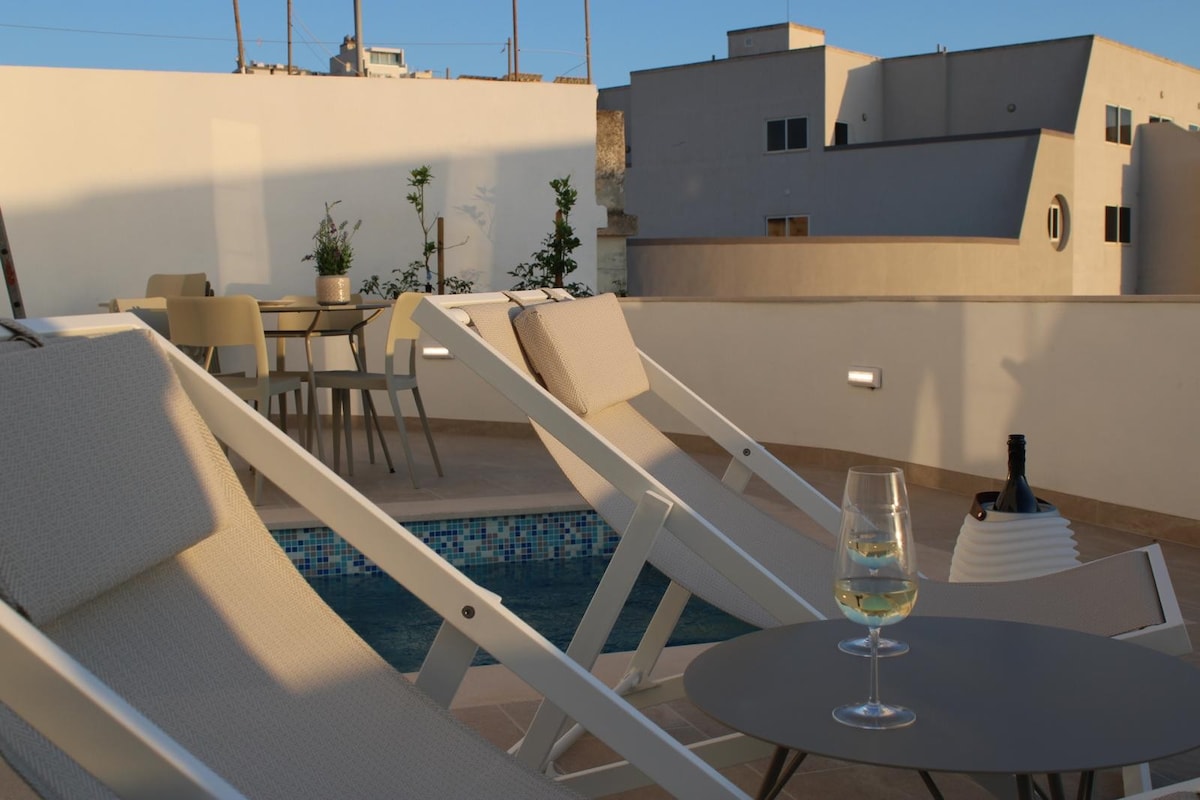A rooftop terrace is shown with two lounge chairs positioned near a small table, which holds a glass of white wine and a bottle. A private pool is visible, surrounded by tiles, with the skyline of nearby buildings in the background.
