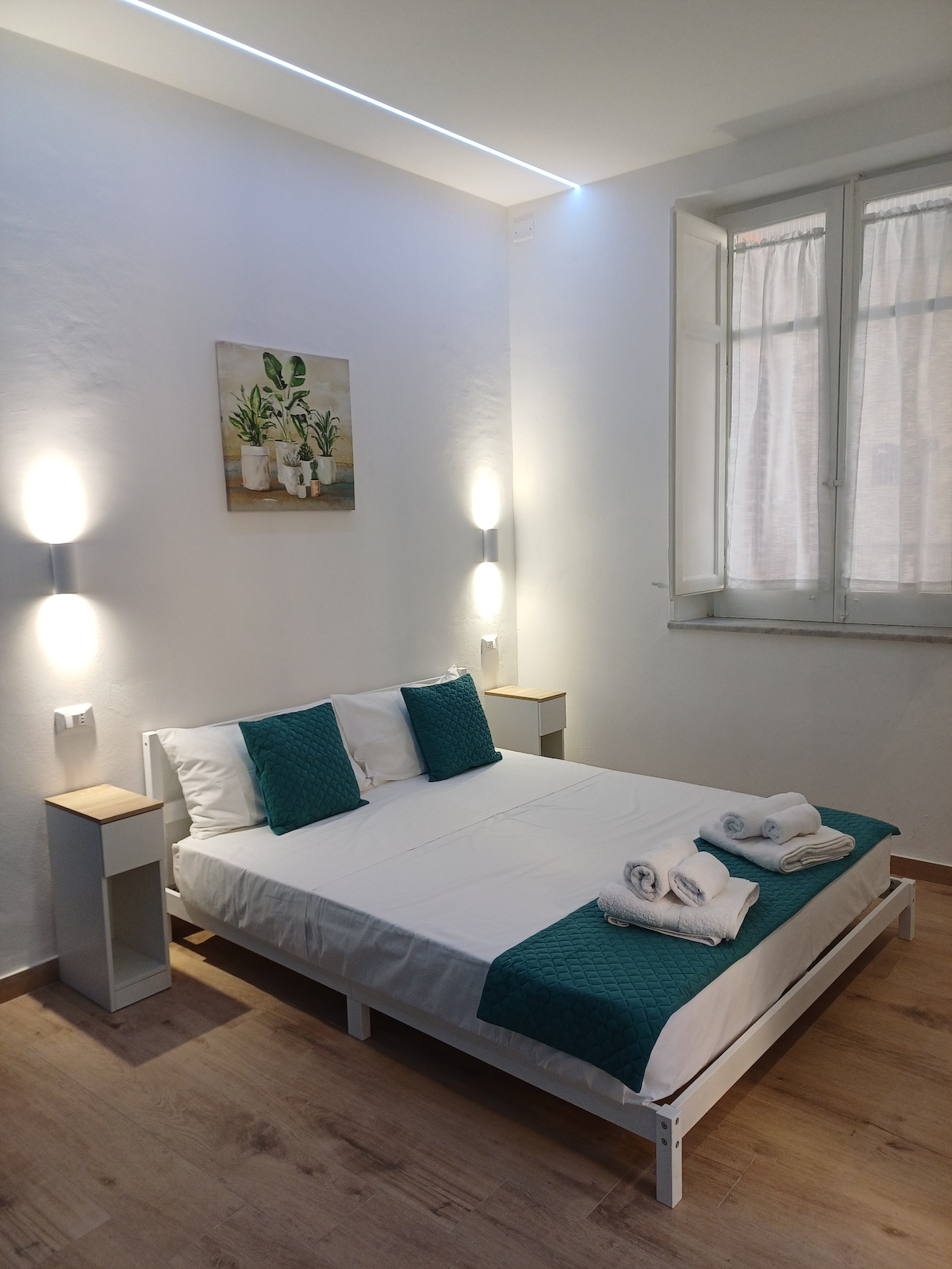A well-lit sleeping area features a spacious bed with turquoise accents, complemented by two bedside tables with lamps. Soft white linens and neatly arranged towels can be seen. Natural light enters through a window, enhancing the minimalist decor of the room.