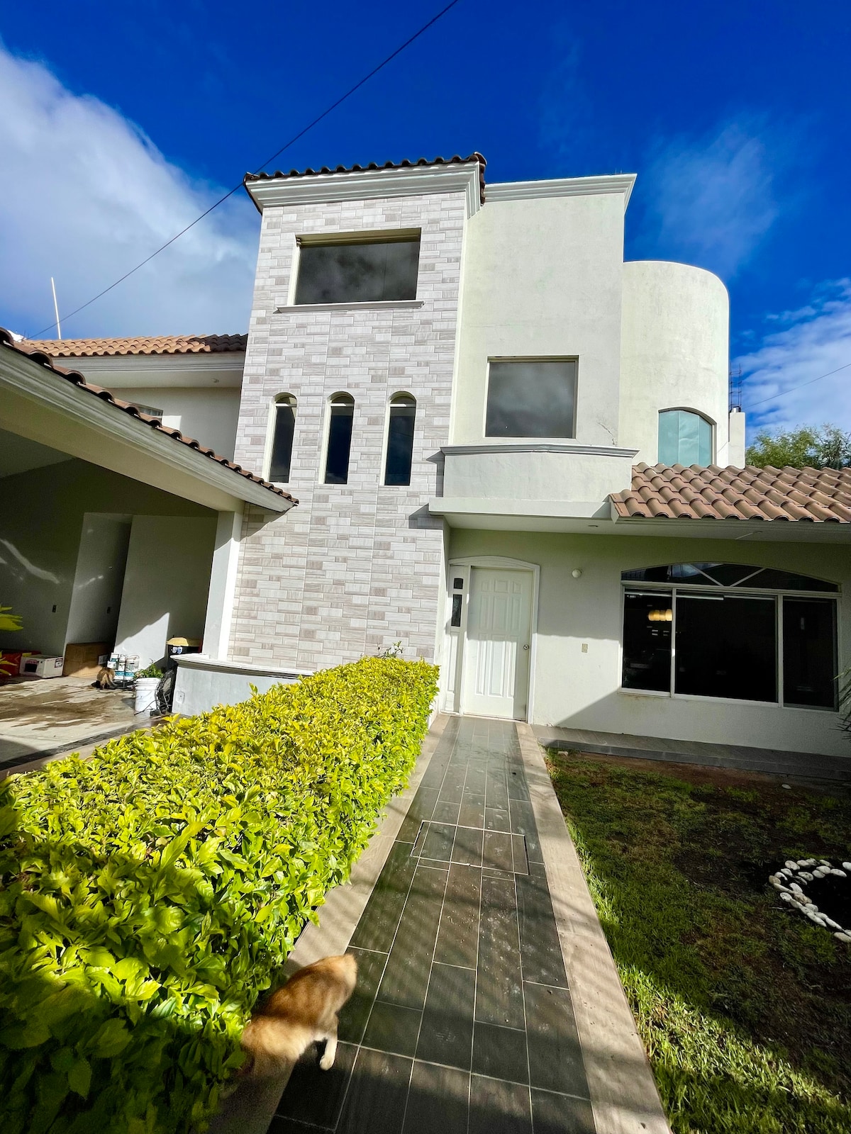 A modern two-story structure features a light-colored exterior with stone accents and large windows. A manicured pathway leads to the entrance, bordered by lush greenery. The roof has a traditional tile design, contrasting with the contemporary facade.