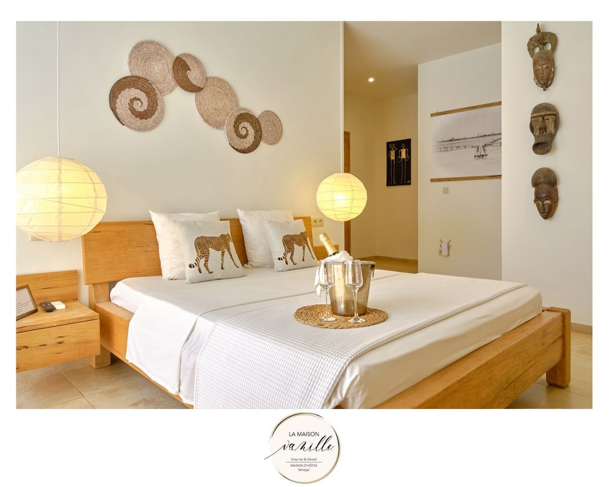 A spacious bedroom is furnished with a wooden bed adorned with patterned pillows. Soft lighting is provided by round lamps, creating a calm ambiance. Decorative wall art, including woven pieces and framed prints, enhances the modern aesthetic of the room.