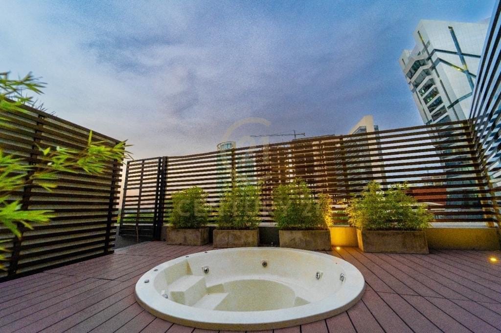 A spacious outdoor area features a circular jacuzzi surrounded by low wood paneling and decorative planters filled with greenery. Soft lighting illuminates the space, creating a serene setting. The view includes nearby buildings under a twilight sky.