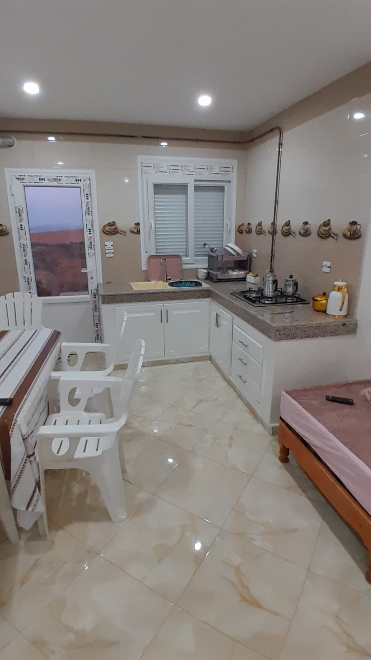 A functional kitchen area is presented with tiled walls and a countertop. A dining table with white plastic chairs is situated beside the window. Kitchen appliances and utensils are neatly arranged on the counter, while warm lighting illuminates the space.