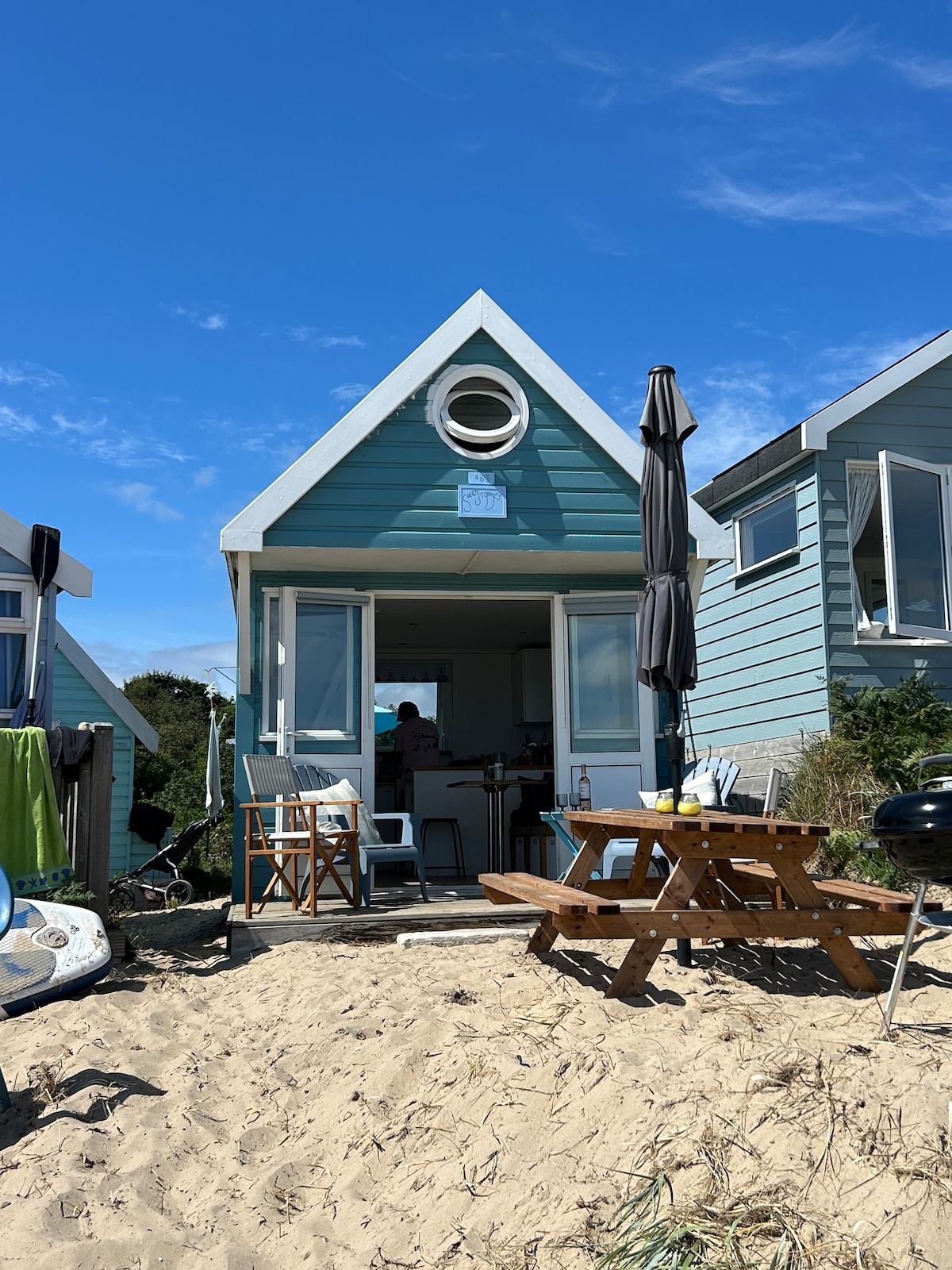 The beach hut features a distinctive sloped roof and large front windows that allow natural light to fill the interior. A picnic table is set outside on the sandy ground, accompanied by an umbrella for shade. The inviting entrance leads into the cozy living space.