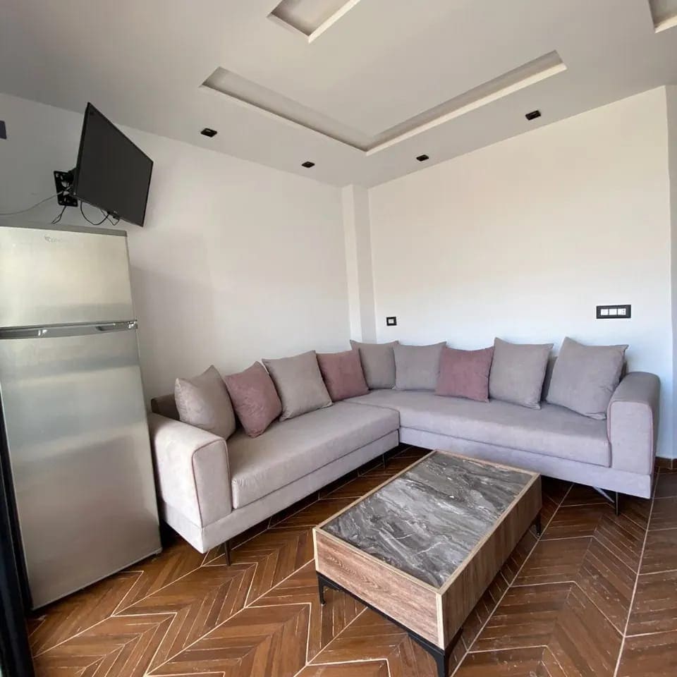 A corner sofa adorned with plush cushions provides ample seating in a well-lit living area. A stylish coffee table with a marbled surface sits in front. A refrigerator is positioned nearby, and a wall-mounted television adds entertainment options.