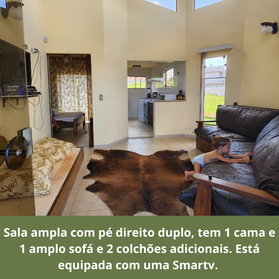 The spacious living area features high ceilings and includes a single bed and a large sofa. A kitchen area is visible in the background, and a cowhide rug lies on the floor. Natural light fills the room through the open doorway.