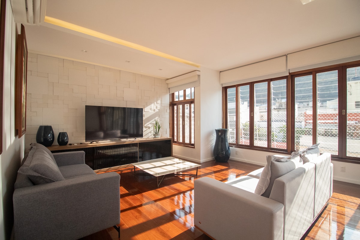 A modern living area is showcased with ample natural light flowing through large windows. Two cozy sofas are positioned around a low coffee table, and a sleek television is mounted on the wall. Decorative elements include a plant and stylish vases, enhancing the space's contemporary feel.