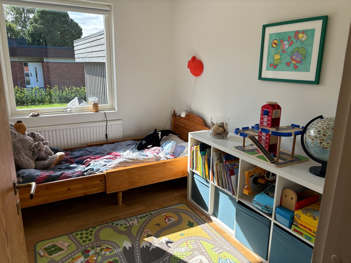 A child's bedroom is illuminated by natural light from a large window. A wooden bed is positioned against one wall, complemented by a playful rug. A storage unit holds colorful toys and books, while a framed artwork adds a cheerful touch to the space.