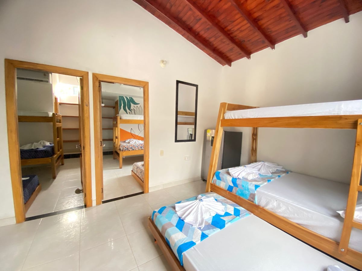 A shared room is highlighted by a wooden bunk bed with spacious mattresses and a tiled floor. A mirror is mounted on the wall, and doorways lead to additional sleeping areas. Natural light fills the space, accentuating the calm atmosphere.