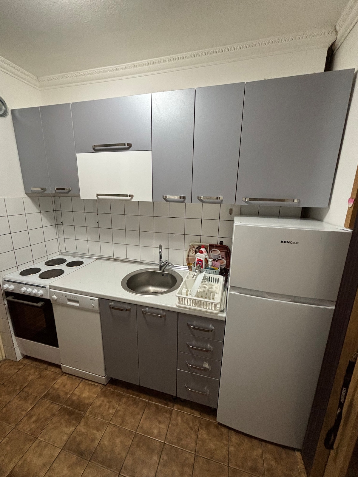 A compact kitchen features modern gray cabinetry and a white countertop. Included are an electric stove, sink, and dishwasher. A refrigerator stands adjacent, and dishware is organized in a nearby drying rack. Bright tiles create a clean and functional space.