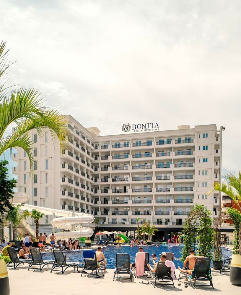 A modern five-story building is visible, featuring multiple balconies and large windows. In the foreground, a pool area with sun loungers is shown, while guests enjoy the water and surroundings. Green palm trees add a tropical touch to the resort ambiance.