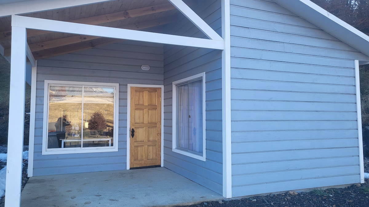 The exterior of the cabin features light blue wooden siding with a peaked roof and white trim. A welcoming wooden door is positioned centrally beneath an overhang. A large window provides natural light, reflecting the outdoor landscape.