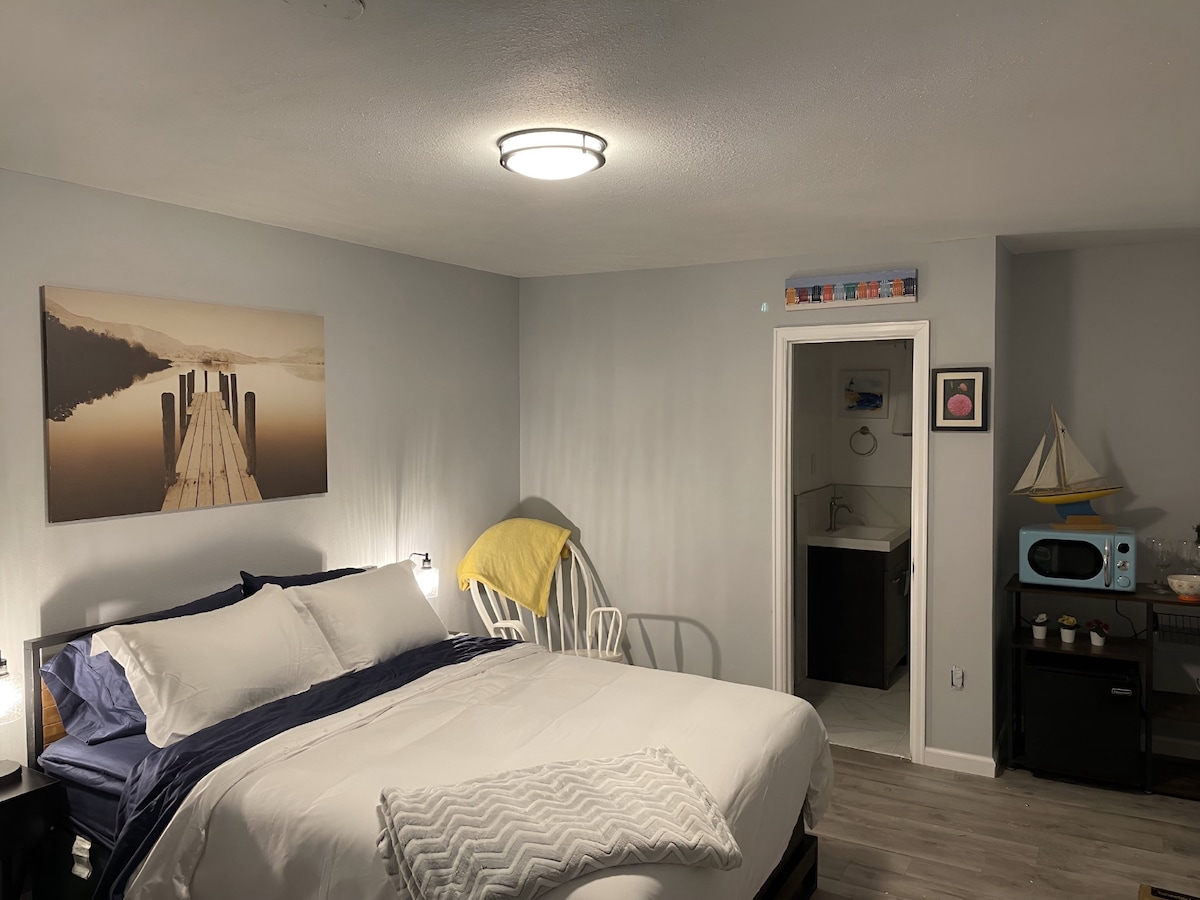 A spacious bedroom features a large bed with crisp white bedding and navy accents. A calming artwork depicting a dock extends across the wall. A yellow blanket adds a pop of color. A doorway on the right leads to a bathroom area, which is partially visible.