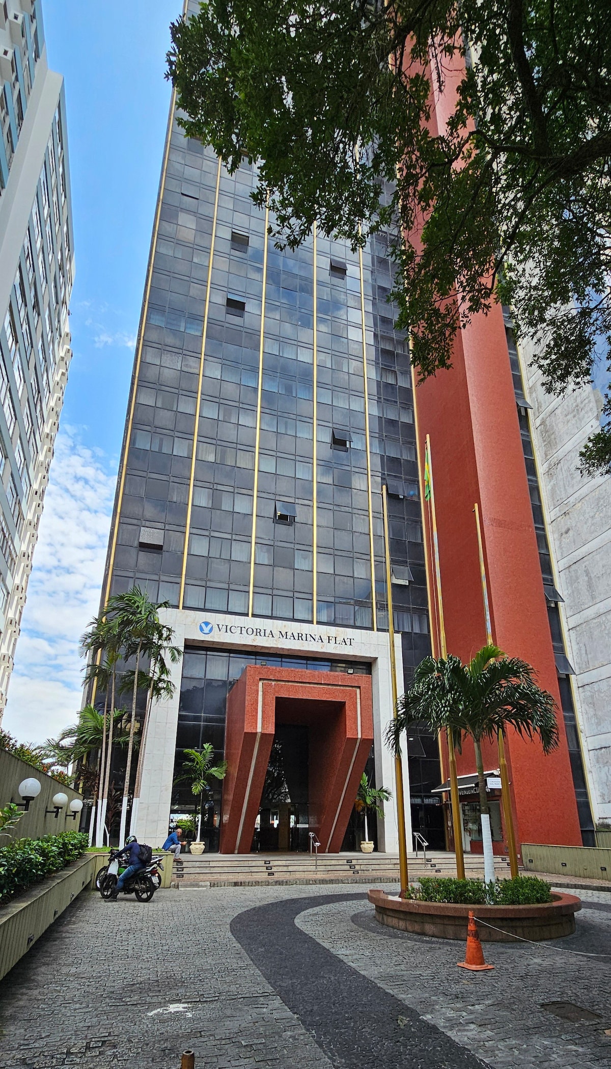 The building features a modern facade with large glass windows and a striking entrance design. Surrounding greenery includes palm trees and landscaped areas. The pathway is paved with cobblestones, leading to the main entrance, while clear skies above provide ample natural light.