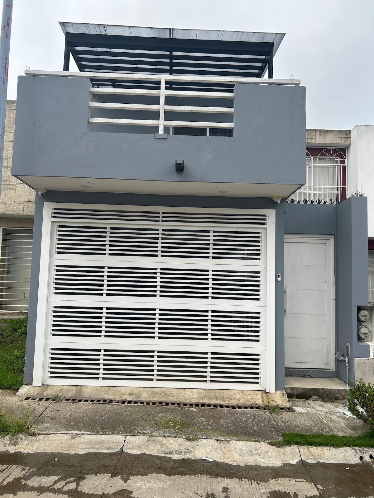 A modern two-story building features a sleek façade in shades of gray. A stainless steel garage door with horizontal slats dominates the ground level, complemented by a metal accent panel and a small entrance door. Lush greenery surrounds the entrance, adding a fresh contrast.