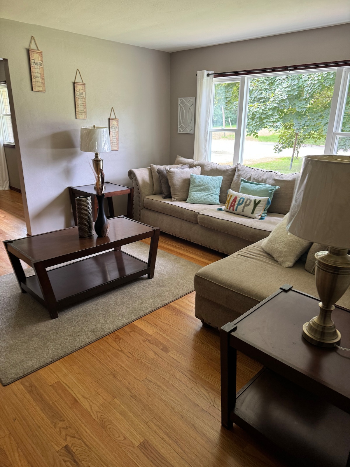 A cozy living room space features a light-colored sofa and loveseat, complemented by decorative pillows. A dark wooden coffee table sits atop a neutral area rug, while two lamps provide soft lighting. Large windows invite natural light, framing a view of greenery outside.