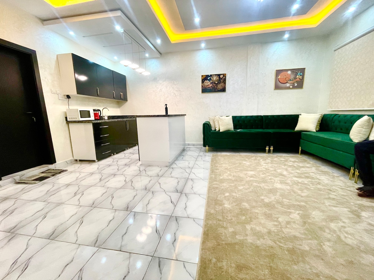 A modern living area is showcased with a green velvet sectional sofa positioned against a light-colored wall. A sleek kitchen is visible in the background, featuring dark cabinetry and a countertop, complemented by stylish lighting fixtures and artwork on the walls.