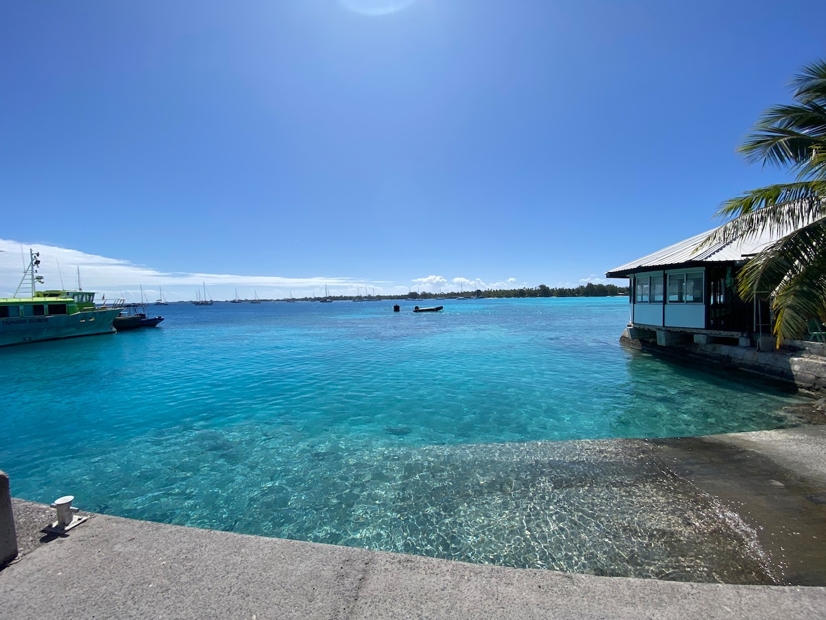 A clear, turquoise lagoon is visible, with gentle waves lapping against the shore. A small boat can be seen in the distance, while a wooden structure with a metal roof is located nearby. Palm leaves frame the right side of the image against a bright blue sky.