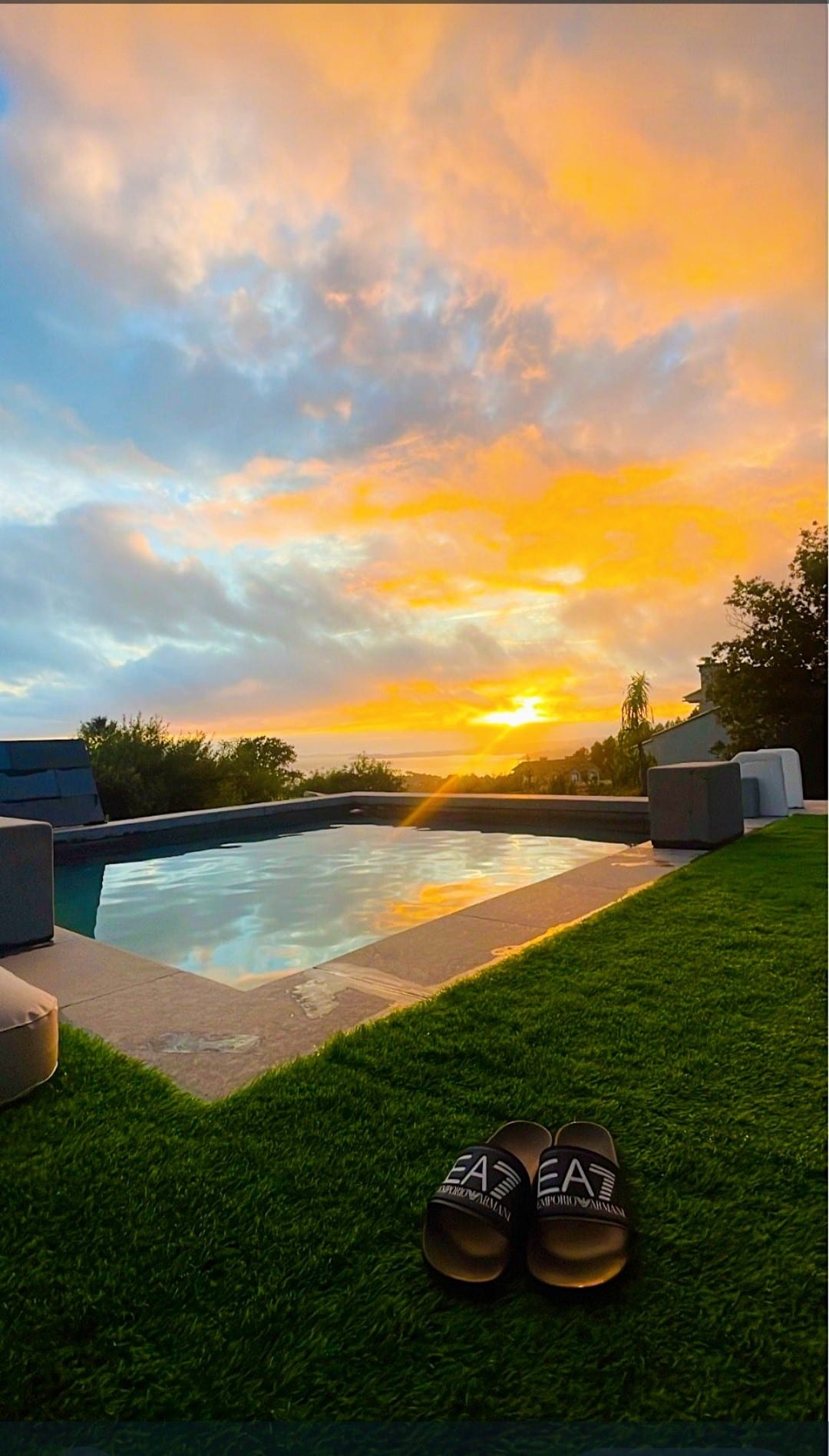 A serene pool is bordered by natural stone tiles and lush green grass, reflecting the vibrant colors of a sunset. Comfortable seating is arranged nearby, and a pair of slides rests on the grass, inviting relaxation in this tranquil outdoor space.