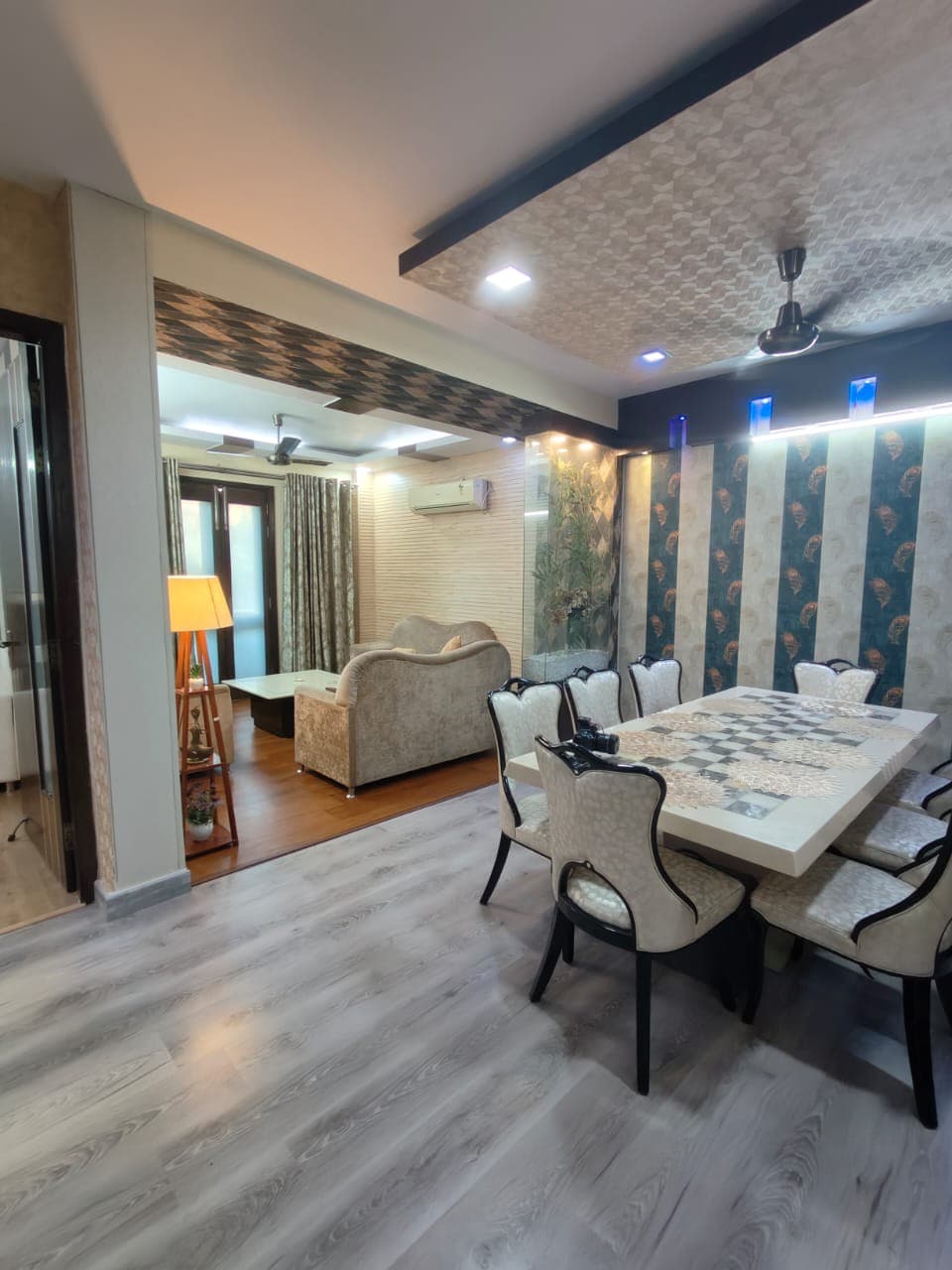 A spacious dining area features a decorative marble table surrounded by six elegantly upholstered chairs. Soft lighting highlights the textured walls, while a cozy seating area can be seen in the background. Natural light filters in through large windows, creating a welcoming environment.