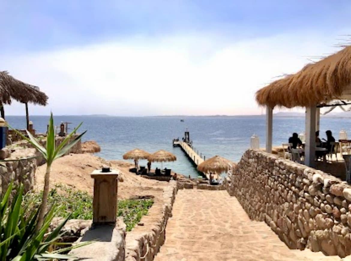 A sandy pathway leads to a wooden pier extending into the calm sea. Thatched umbrellas provide shade on the beach, while natural stone walls frame the scenic view. Soft waves can be seen lapping at the shoreline under a clear blue sky.