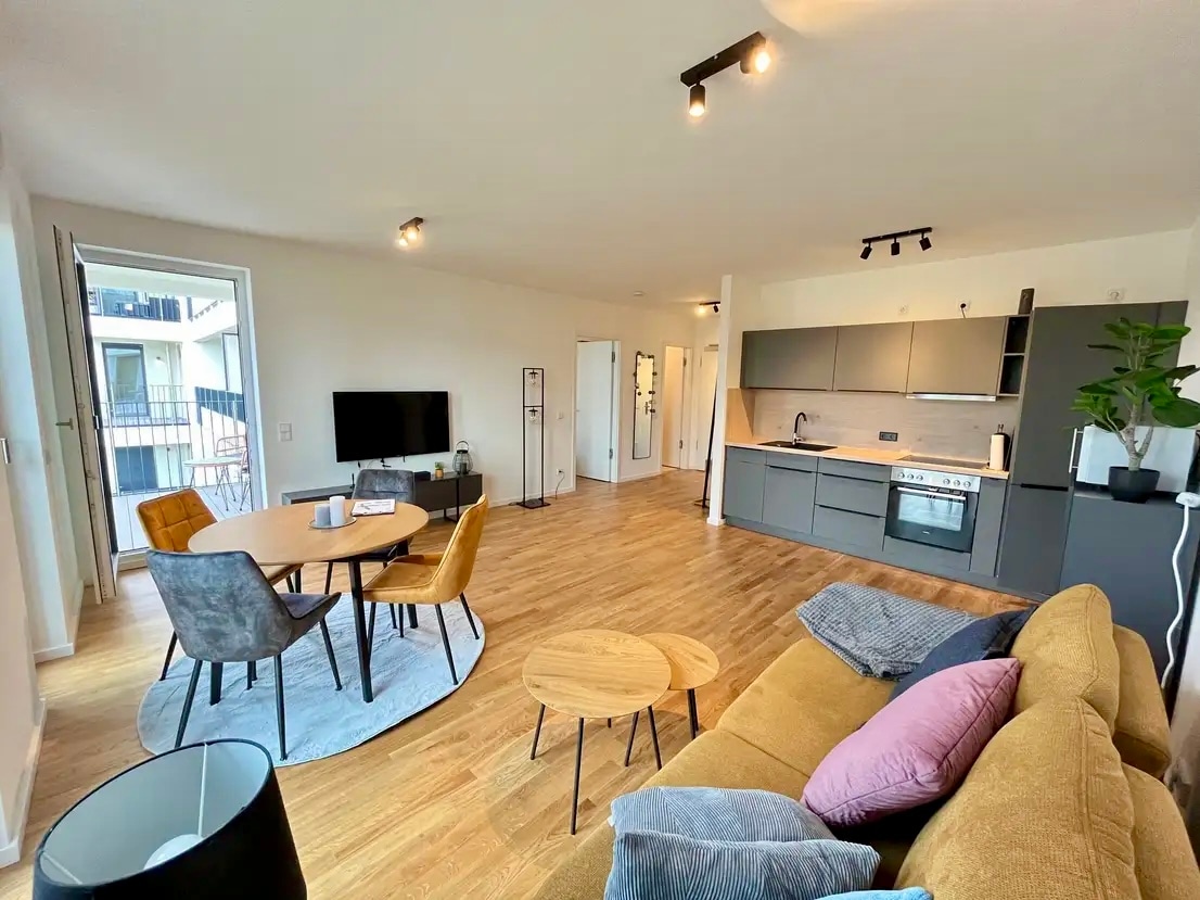 An open living space is depicted, featuring a cozy yellow sofa, a round dining table with four chairs, and a modern kitchen area equipped with a stove and oven. A television is placed on a stand, with natural light streaming through large windows, highlighting the wooden floor.
