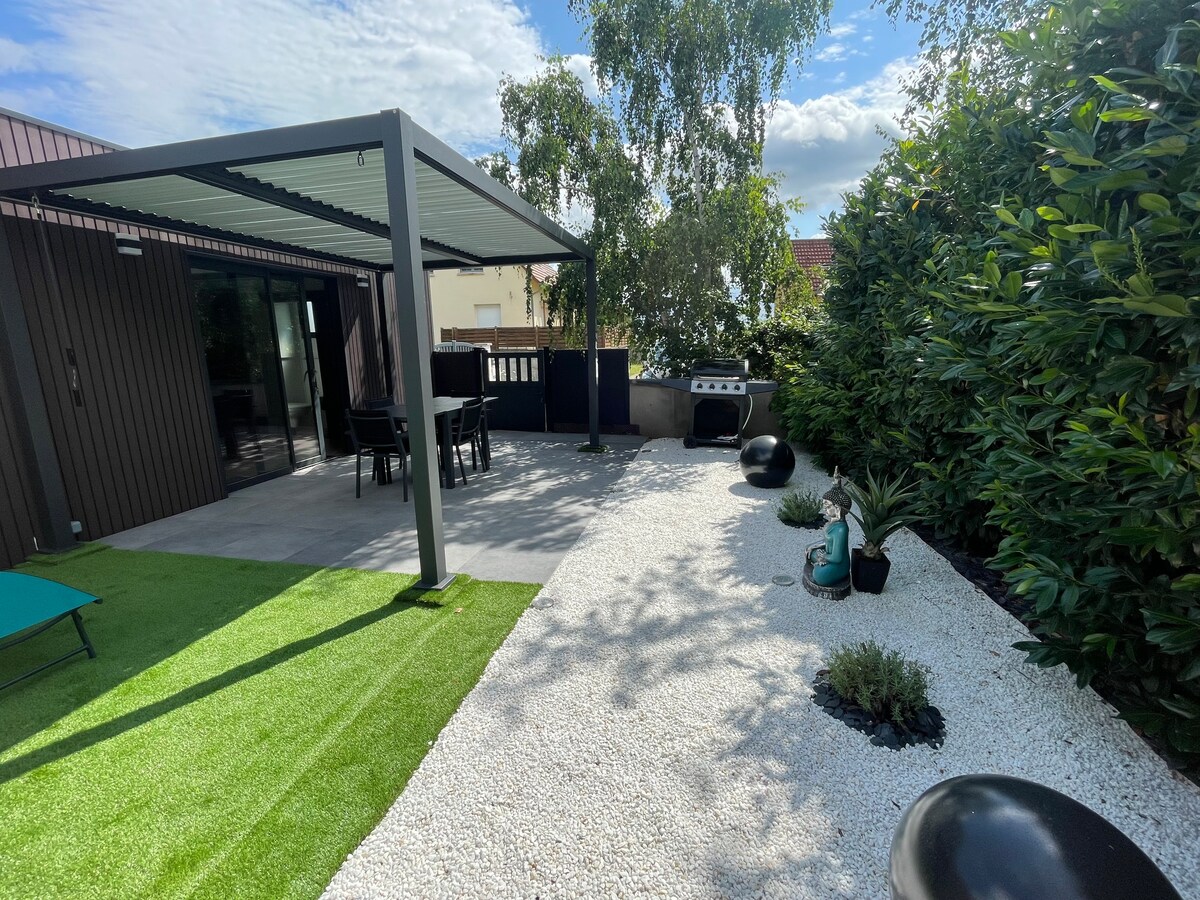 An outdoor patio area is presented under a modern pergola. A dining table and chairs are arranged for al fresco meals, while a gas barbecue sits nearby. The space features well-maintained gravel landscaping, artificial grass, and potted plants providing greenery.