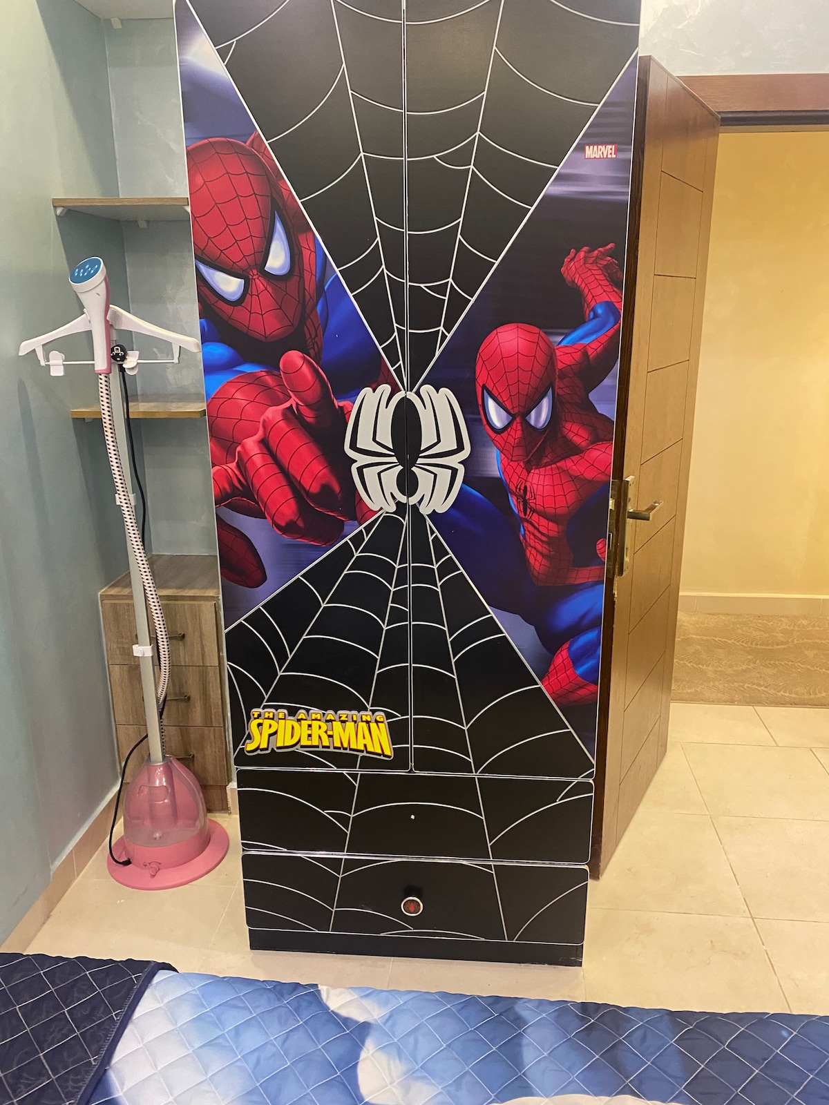 A colorful wardrobe featuring vibrant illustrations of Spider-Man, depicted in dynamic poses against a black background embellished with web patterns. The wardrobe stands in a well-lit space with neutral-colored walls and tiled flooring, creating a playful focal point within the room.